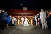 Bride and groom walking hand in hand out of a red-roofed pavilion at night as guests line both sides and sparkler fountains glow behind them
