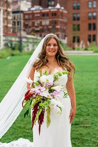 Smiling bride on a city lawn holding a bold white, pink and deep red bouquet, her veil trailing beside her