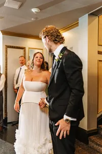 Smiling bride in a strapless gown holding the groom's hand as guests look on