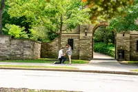 Man on one knee holding out a ring as she smiles down at him beside an old stone gateway in Frick Park, Pittsburgh
