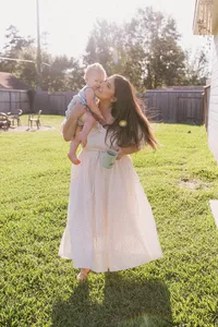 A mother dressed in a white dress holds her coffee in one hand and her toddler in another in her backyard during their at home family photo session near houston texas