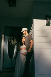 A husband and wife stand in the sunshine of their front porch during their maternity photos in houston texas