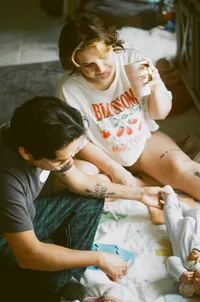 A film photo of a mother and father enjoying their baby laying on a blanket while the mother drinks coffee during their at home family photo session near houston texas