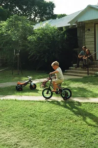 A film photo of a boy riding his bicycle in the sunshine while his parents watch in their front yard during their at home family photo session near houston texas
