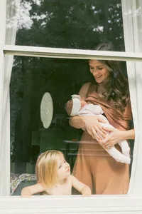 A mother holds her newborn and smiles down at her toddler through then window of her home during her in home newborn photo session in houston tx