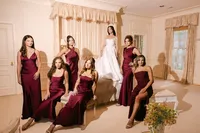 Editorial portrait of bride Amanda standing on a bed surrounded by her bridesmaids in burgundy satin dresses, by Dove and Fox