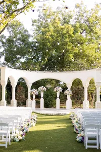 An outdoor wedding venue with white chairs arranged in rows, floral decorations along the aisle, string lights overhead, and a white archway in front, surrounded by trees.
