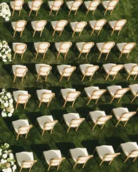 white wooden chairs on lawn - Photograph of rows of white wooden ceremony chairs arranged on green grass with white floral arrangements lining the aisle