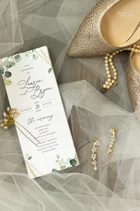 Wedding invitation with floral details, pearl jewelry, gold earrings, and beige high heels arranged on white tulle fabric.