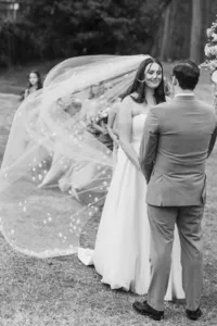 Bride and groom facing each other as her long lace-trimmed veil blows in the wind