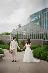 A bride and groom hold hands as they walk towards the camera, a glass greenhouse behind them.