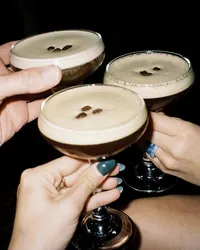 three espresso martinis being held close together as if "cheersing" and three white hands holding the glasses