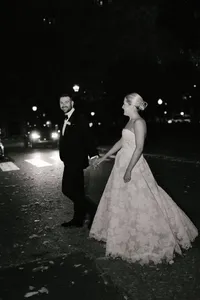 Bride and groom walk hand in hand down a city street lit by car headlights at night