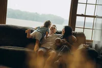 Candid indoor family photo of a mother and father playing with their toddler on the couch as they hold their newborn.