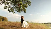 Wedding videography still of couple standing together in open grassy field with trees and sky backdrop