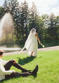 bride by lakeside with tall trees - Photograph of bride in white gown standing on grass near still water with tall evergreen trees reflected and groom visible in background