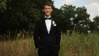 Wedding videography still of groom in formal black tuxedo standing alone in outdoor grassy area with trees