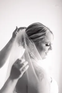 A bride smiles gently as someone adjusts her veil. The image is in black and white, capturing a soft, intimate moment before the wedding ceremony.