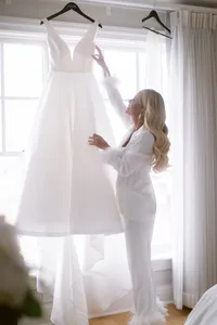 Bride in a white robe reaches up to admire her wedding gown hanging by a bright window