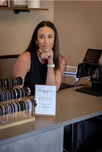 Woman jewelry designer smiles behind a display of colorful bracelets, with a handwritten sign and podcast microphone on the studio table.