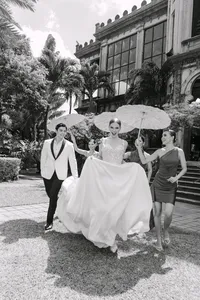 Black-and-white photo of a bride and groom with friends holding parasols in front of a grand mansion