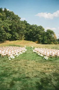 outdoor ceremony aisle setup - Photograph of outdoor ceremony space with white wooden chairs arranged in rows down center aisle on manicured lawn with trees