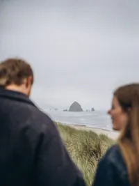 A man and woman look at each other, but they're blurred so that Haystack Rock in the background is the focus.