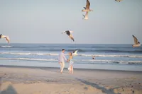 Couple walking the beach as birds fly overhead during their engagement session