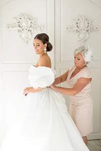 A smiling bride in an off-shoulder white gown stands as an older woman in a light pink dress helps adjust her dress. They are in a bright, elegant room with white ornate decor on the wall.