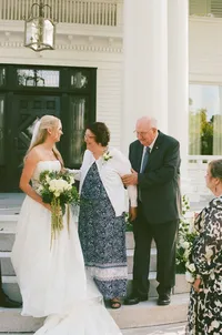 Photograph of bride in white gown greeting family members at formal entrance with ivy-covered green door