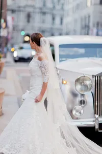 Bride in a long sleeve lace gown stands beside a vintage Rolls-Royce on a city street