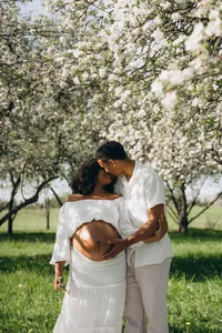 A man holds his pregnant wife's belly as they smile at each other in an orchard of trees with white flowers around them.