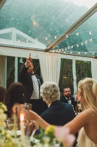 reception guests under tent with string lights - Photograph of bride holding bouquet surrounded by guests gathered under marquee tent with overhead string lights
