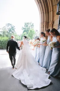 Bride and groom walk hand in hand past bridesmaids in pale blue gowns under a stone archway