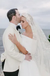 Groom kisses bride's forehead by the ocean as her lace veil blows in the wind