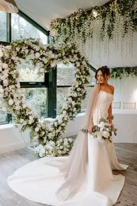 bride with a white flower bouquet in front of a heart-shaped white rose wedding arch