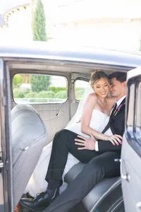 A bride and groom sit closely together in the back seat of a vintage car, smiling and embracing, dressed in wedding attire. Sunlight fills the car, highlighting their happiness.