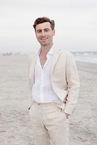 Groom in a tan linen suit smiles on the beach with the ocean behind him