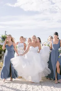 Bride laughs while walking on the beach surrounded by bridesmaids in dusty blue gowns