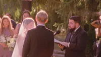 Wedding videography still of guests in formal attire at outdoor ceremony with floral arch