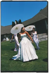 A bride smiles as 2 of her bridesmaids walk behind her, all holding white parasols.