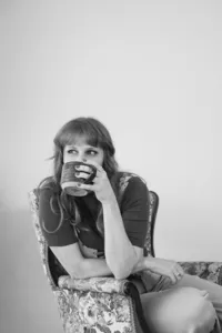Black-and-white photo of a woman with bangs sitting on a patterned chair, holding a mug with both hands near her mouth and looking to the side, appearing thoughtful. The background is plain.