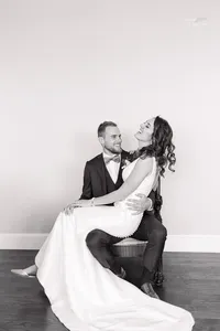 A bride in a long gown sits on a grooms lap. Both are smiling and dressed formally. They pose against a plain wall in a light, airy room with wooden floors.