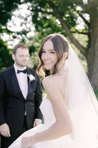 Bride glances back at the camera smiling as her groom stands beneath a shaded tree canopy