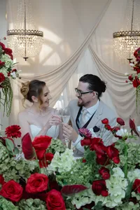 wedding couple clinking glasses surrounded by red and burgundy floral arrangements