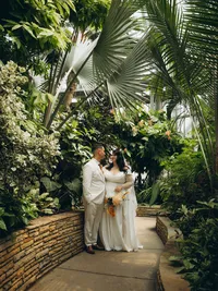 A bride and groom smile at one another as they stand inside of a lush greenhouse, by Detroit wedding photographer Grace Wan.