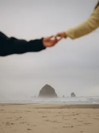 Close up of 2 hands reaching out touching each other, blurred so that Haystack Rock is the focus in the background.