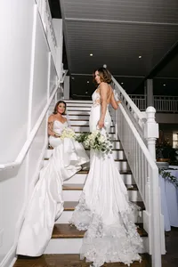 Brides Chyan and Allison in white gowns holding bouquets on a staircase at their wedding venue, by Dove and Fox