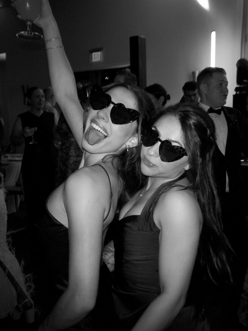 Two friends having fun in heart-shaped sunglasses at a wedding