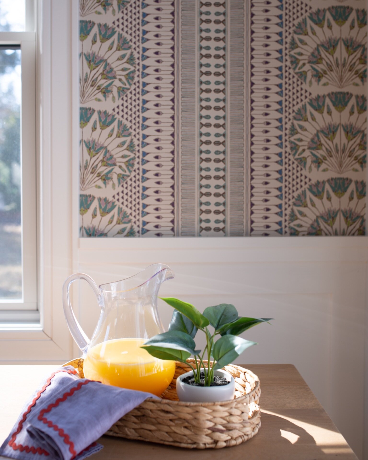 Breakfast nook with patterned wallpaper, a glass pitcher of orange juice, and a potted plant on a woven tray